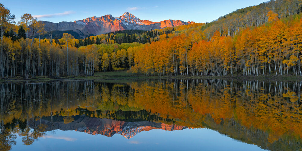 Wilson's Mirror Panoramic landscape photograph of Mount Wilson glowing in warm sunrise light above golden autumn aspen trees, perfectly reflected in a calm alpine pond in Color